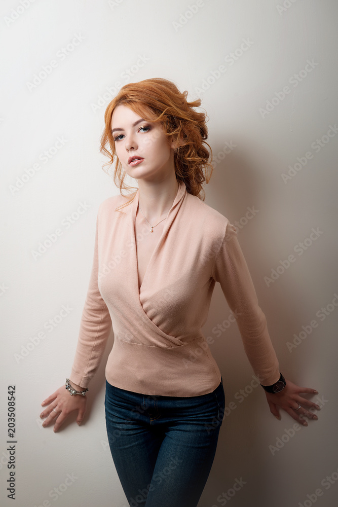 Red-haired girl in jeans and a pink blouse posing in a fashion style on a white isolated wall background. Red-haired girl in jeans and a pink blouse posing in a fashion style on a white isolated wall background.