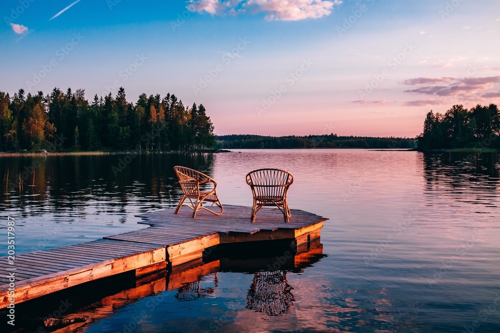 Two wooden chairs on a wood pier overlooking a lake at sunset - obrazy, fototapety, plakaty 