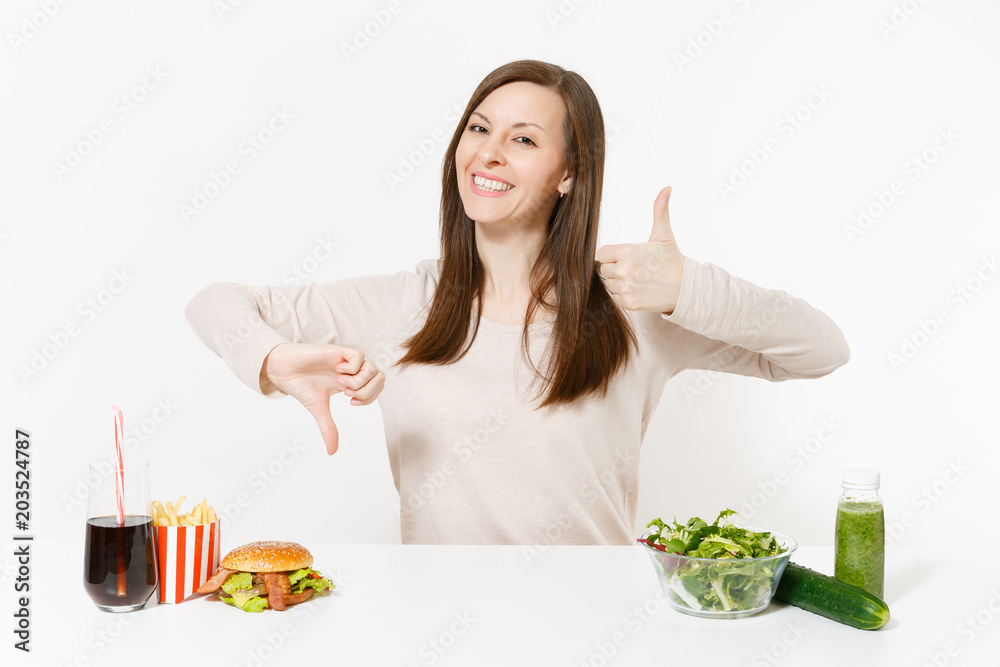 Woman shows thumbs up down with green detox smoothies, salad in glass bowl, cucumber burger cola in bottle isolated on white background. Proper nutrition, healthy lifestyle, fast food, choice concept. Woman shows thumbs up down with green detox smoothies, salad in glass bowl, cucumber burger cola in bottle isolated on white background. Proper nutrition, healthy lifestyle, fast food, choice concept.