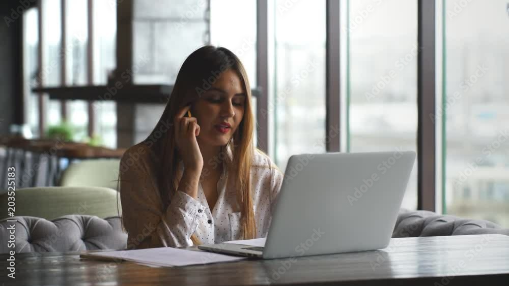 Beautiful young woman working with laptop and talking on smartphone talking to customer sitting near window