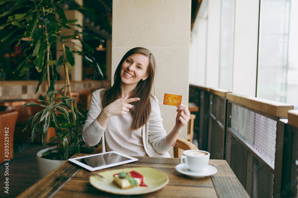Woman sitting near big window in coffee shop at table with credit card, cup of coffee cake relaxing in restaurant during free time. Female working on tablet pc computer rest in cafe. Lifestyle concept Woman sitting near big window in coffee shop at table with credit card, cup of coffee cake relaxing in restaurant during free time. Female working on tablet pc computer rest in cafe. Lifestyle concept