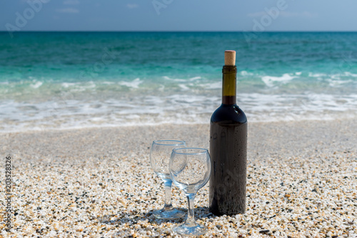 Bottle of red wine with wine glasses on the beach at the summer sunny day. Sea on the background.