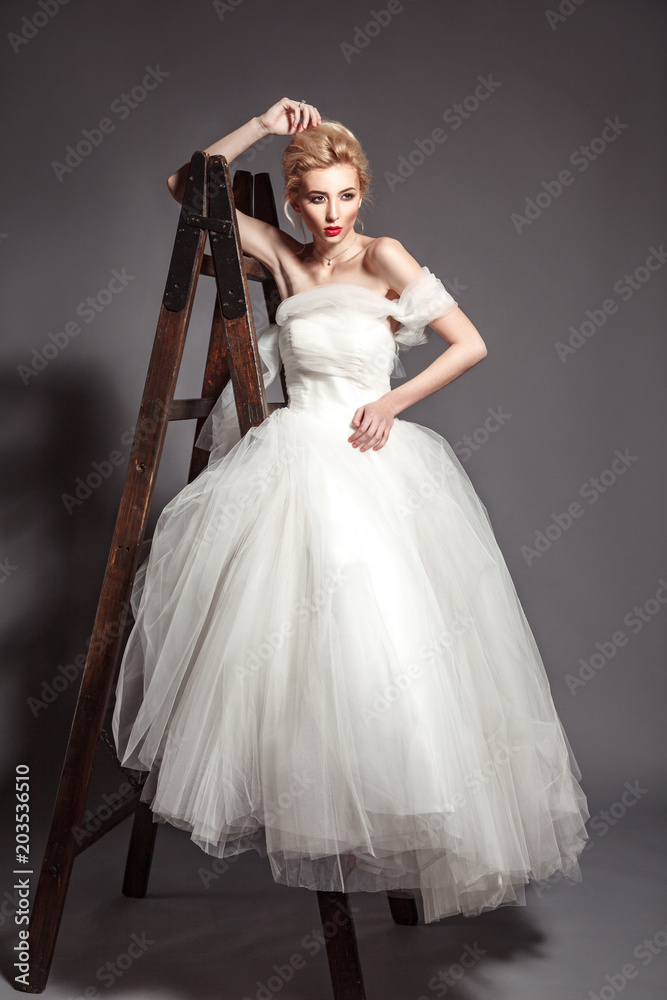 Fototapeta premium portrait in vogue style of fashion beautiful bride in wedding dress leaning on wooden ladder on grey background