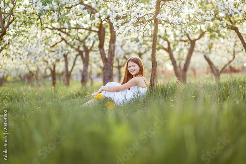 A portrait of a beautiful young girl with blue eyes in a white dress  with dandelions in the garden with apple trees blosoming having fun and enjoying smell of flowering spring garden at the sunset