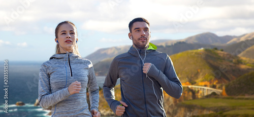 fitness, sport and technology concept - happy couple running and listening to music in earphones over bixby creek bridge on big sur coast of california background
