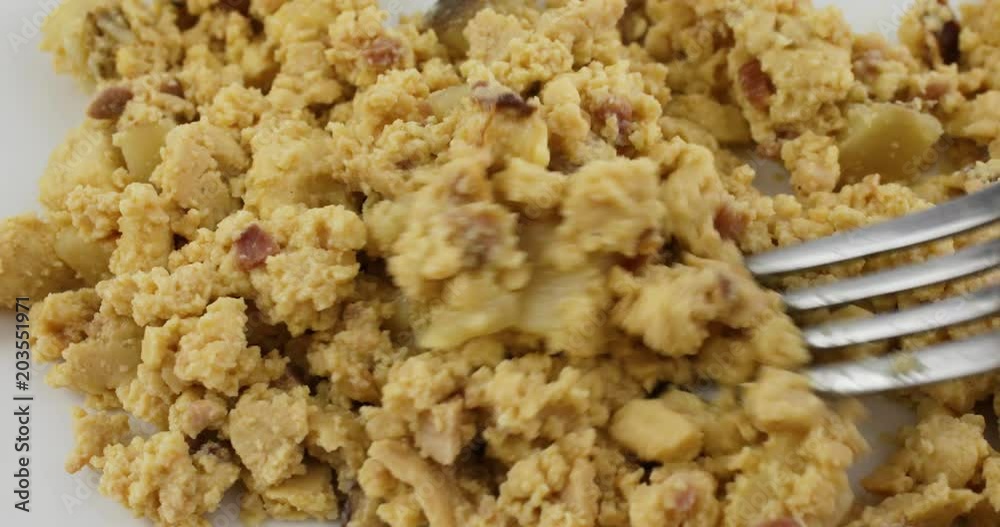 Close view of a meal of scrambled eggs with potatoes and bacon on a white plate being mixed then taking a forkful at the end.