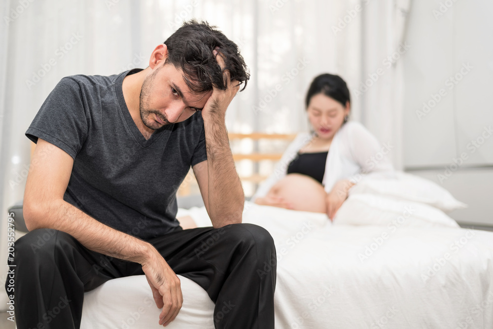 Close Up Of Serious Man Sitting On Bed With Blur Of Unhappy Pregnant Woman With Big Belly.Needless Pregnancy And Motherhood Moment People And Expectation. Family planning Concept.In Selective Focus. Close Up Of Serious Man Sitting On Bed With Blur Of Unhappy Pregnant Woman With Big Belly.Needless Pregnancy And Motherhood Moment People And Expectation. Family planning Concept.In Selective Focus.