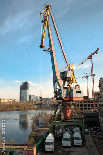 Loading-unloading crane in the river dock of an industrial enterprise. Nearby in the parking lot are three concrete mixer cars. Against the background of the blue sky and the surface of the river.