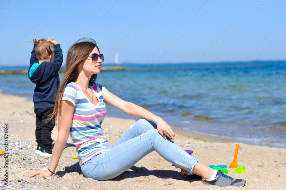 Fototapeta premium Beautiful young woman in sunglasses sits on the beach in the sand and looks into the distance and the child is playing nearby and looks at the sea 