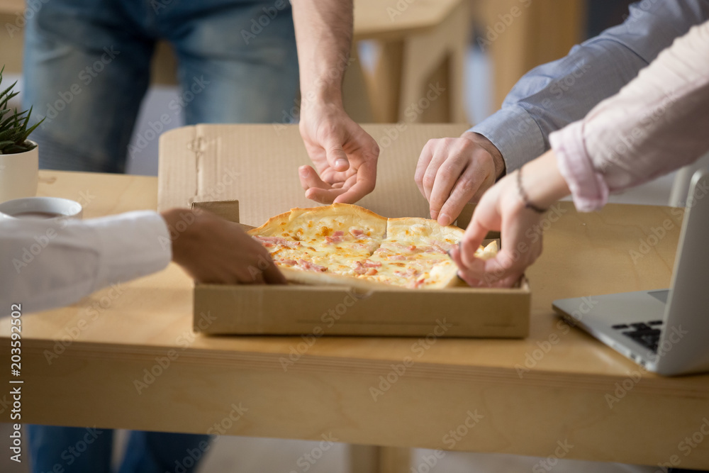 Hands of multiracial people taking cheese and ham pizza slices from box on work desk, employees eat lunch meal together enjoy italian food at workplace, office delivery service concept, close up view