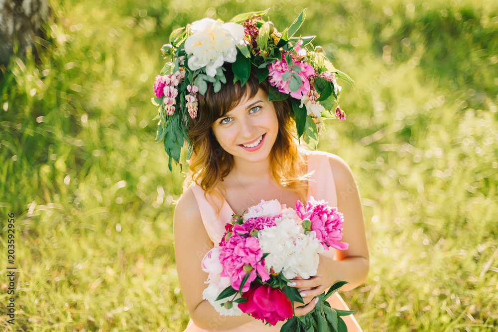 Fototapeta premium portrait of a happy smiling girl with a wreath of flowers and a bouquet of peonies in nature on a Sunny summer day
