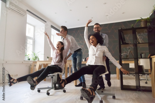 Millennial multiracial team people having fun riding on chairs in office room, excited diverse employees laughing enjoying funny activity at work break, creative friendly workers play game together