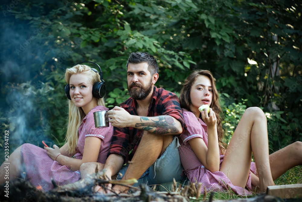 Group of friends sitting next to campfire. Girls leaning on their male friend. Blond woman listening to music while brunette lady eats apple. Smiling guy drinking tea or coffee from mug Group of friends sitting next to campfire. Girls leaning on their male friend. Blond woman listening to music while brunette lady eats apple. Smiling guy drinking tea or coffee from mug