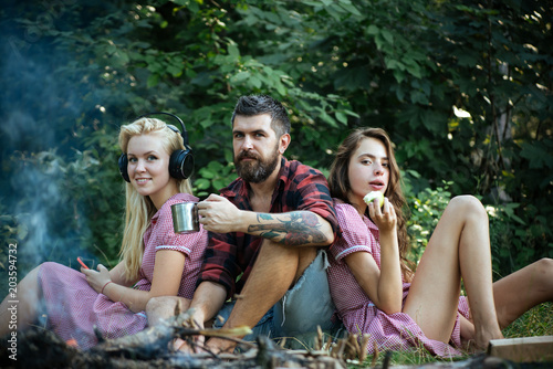 Group of friends sitting next to campfire. Girls leaning on their male friend. Blond woman listening to music while brunette lady eats apple. Smiling guy drinking tea or coffee from mug
