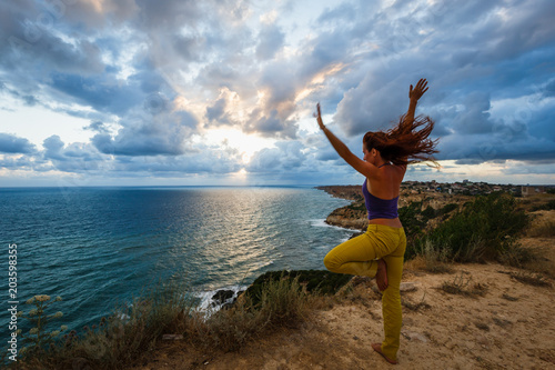 Amazing sunset on the sea. Cloudy sky. A woman is practicing yoga in nature on a cliff on a rocky ocean shore. Wind in the hair. Spirit of freedom. Meditation. Wildness. Wandering. Journey. Pose tree.