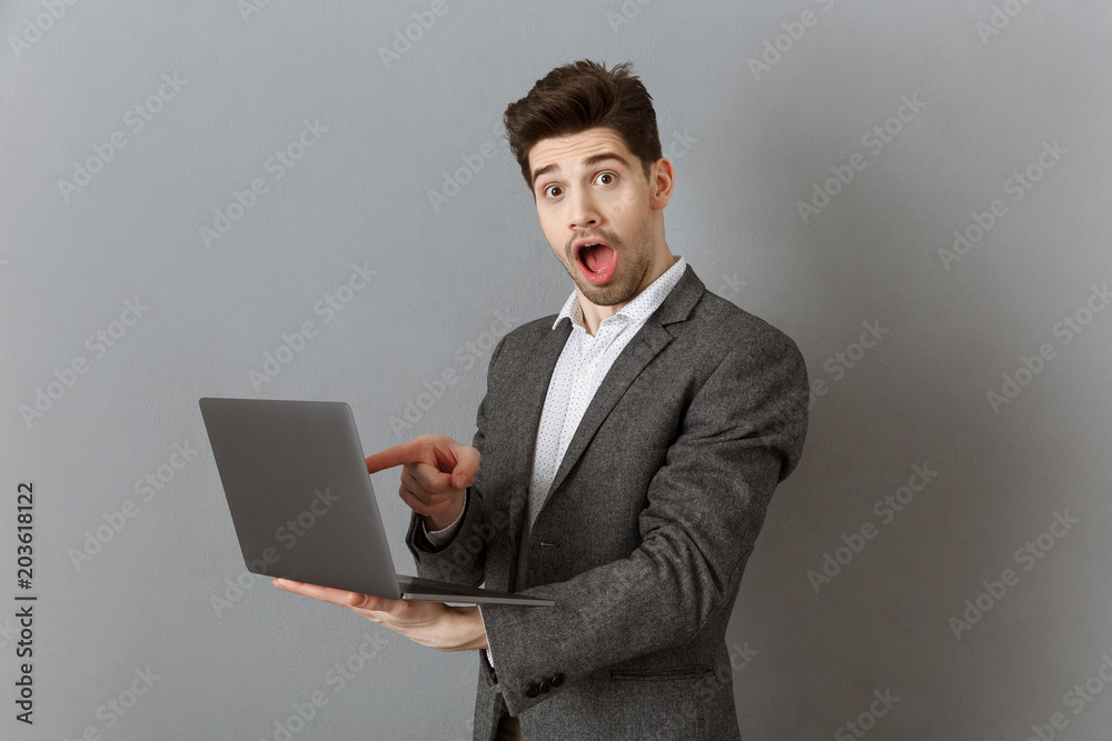 shocked businessman in suit pointing at laptop against grey wall background