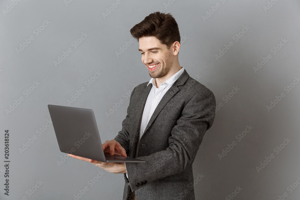 smiling businessman in suit using laptop against grey wall background smiling businessman in suit using laptop against grey wall background