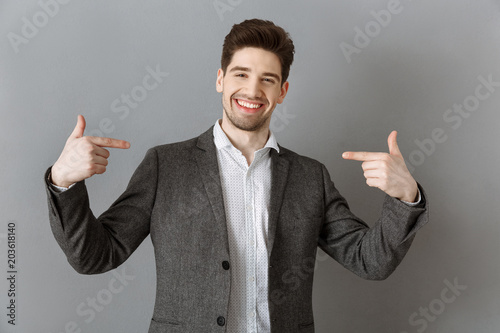 portrait of smiling businessman in suit pointing at himself against grey wall