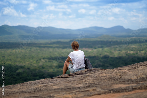 Young woman with backpack sitting on mountain and looking to a sky with clouds. Beautiful nature of Sri Lanka