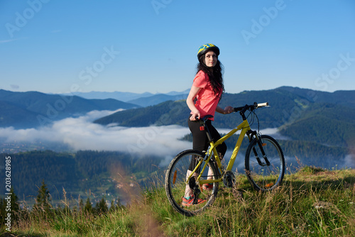 Beautiful female biker with yellow bicycle in the mountains in the morning, wearing helmet and red red t-shirt. Foggy mountains, forests on the blurred background. Outdoor sport activity