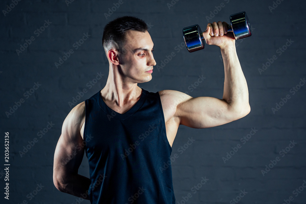 young man strengthens the muscles of the hands of dumbbells in the gym on a black background