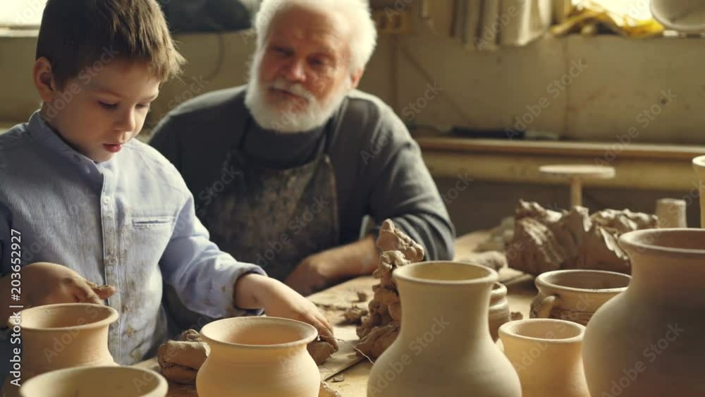 Little boy is playing with clay sitting with his grandparent at table in home pottery studio. Grandfather is talking to curious child, boy is enjoying new hobby. Little boy is playing with clay sitting with his grandparent at table in home pottery studio. Grandfather is talking to curious child, boy is enjoying new hobby.