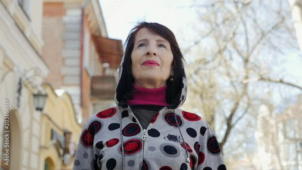 Portrait of a cheery middle-aged woman in a sportive hoodie strolling along an alley with houses in a positive way in spring in slow motion Portrait of a cheery middle-aged woman in a sportive hoodie strolling along an alley with houses in a positive way in spring in slow motion