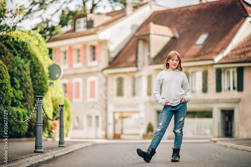 Outdoor portrait of funny little girl wearing grey sweatshirt, denim jeans and black boots