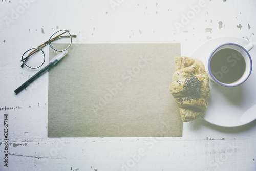 .Creative work concept.Mock-up blank craft sheet of empty paper, pen, eye glasses and morning coffee cup with croissant on white wooden desk. Business background for letter writing.Flares effect photo