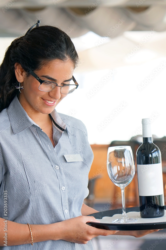 A young waitress carries a bottle of red wine with wine glasses on a tray to the customer of the hotel restaurant. The concept of service. A young waitress carries a bottle of red wine with wine glasses on a tray to the customer of the hotel restaurant. The concept of service.
