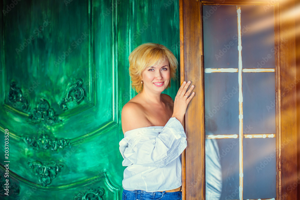 Fototapeta premium Portrait of blond woman in white shirt with bared shoulders standing at door in background of green wall