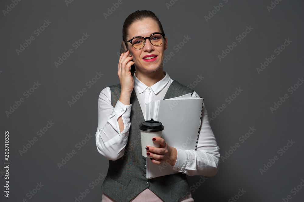 Beautiful young businesswoman in formal suit talking on smartphone and coffee mug standing against gray background Beautiful young businesswoman in formal suit talking on smartphone and coffee mug standing against gray background