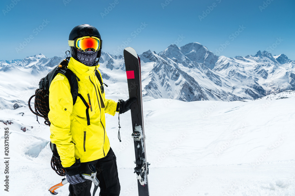 Close-up portrait of a skier wearing a protective helmet and glasses. A mask and scarf with an ice ax in his hands next to skis on the snow-capped mountains of the Caucasus. Skiing