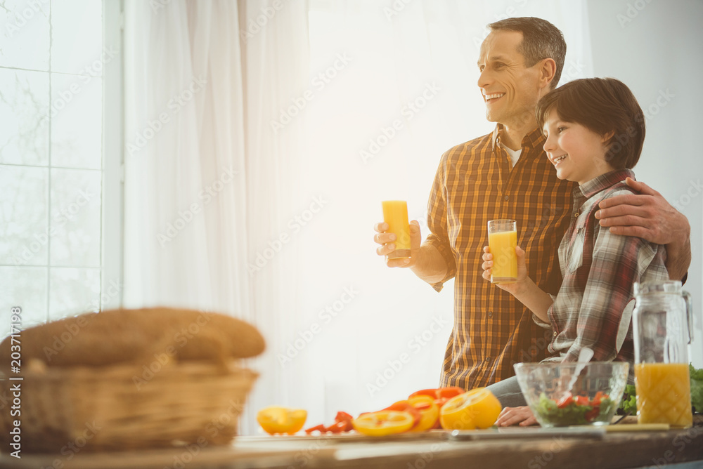 Happy father and kid standing in kitchen in embrace. They holding squeezed juice in hands. Copy space in left side