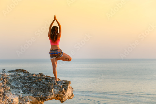 Meditation on the beach. A woman is standing on a cliff above the ocean and doing yoga. Femininity. Force. Soft shades at sunset. Beautiful scenery. You're on top of the world. © mishamif
