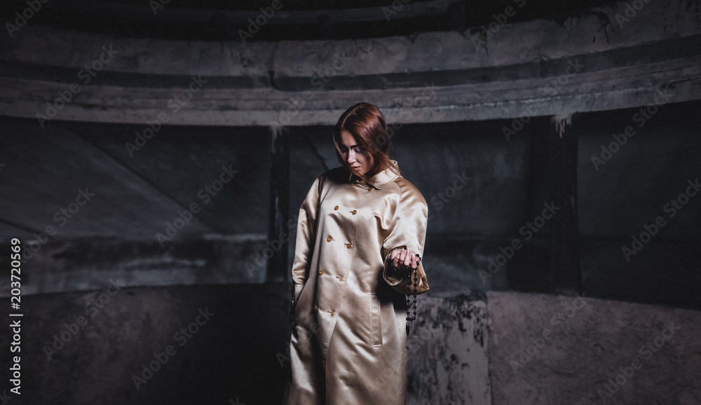 Abstract photo of a beautiful young woman in a beige silk coat against a background of concrete walls of an underground city station with cherries in hand.