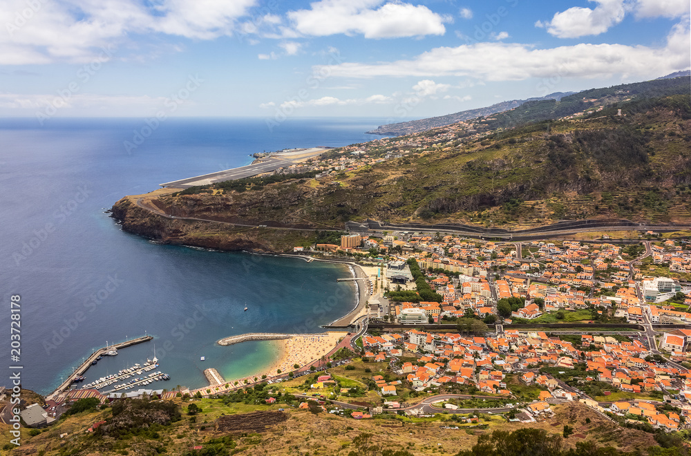View of Machico from the viewpoint of Pico do Facho on a summer day, with the sea on the left side and in the background the Cristiano Ronaldo Airport of Madeira, Portugal.