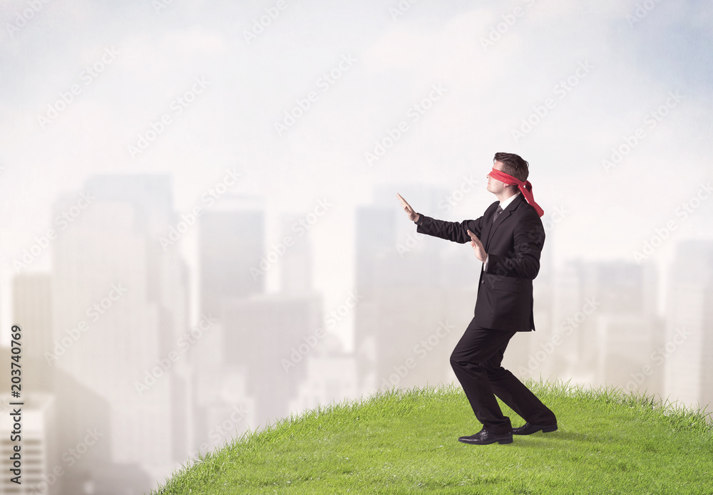 Young blindfolded businessman steps on a a patch of grass with a city in the background Young blindfolded businessman steps on a a patch of grass with a city in the background