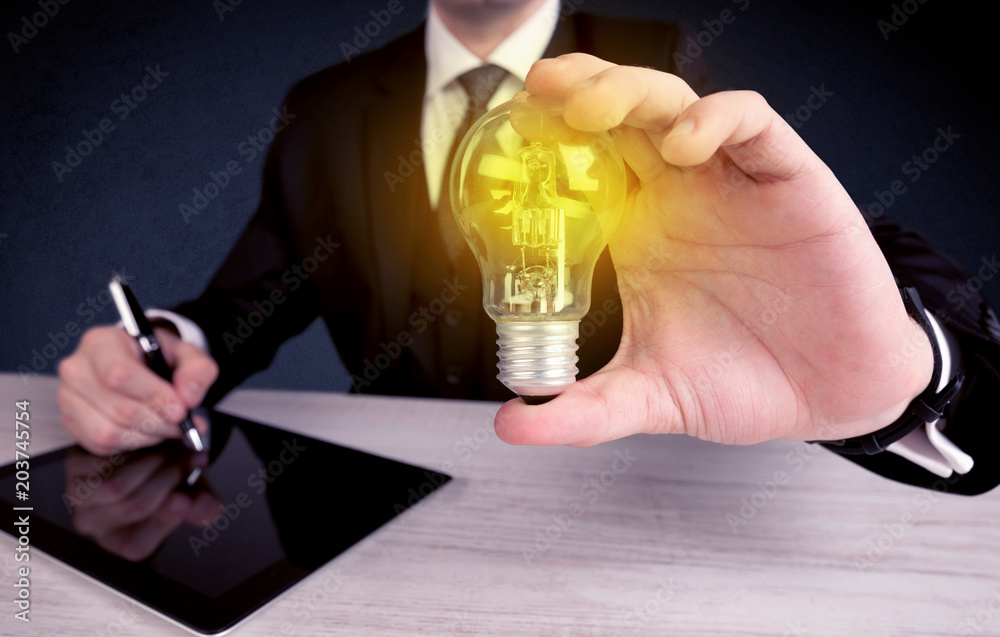 A business man in suit holding a glowing yellow light bulb in his hand while working in the office concept. - obrazy, fototapety, plakaty 
