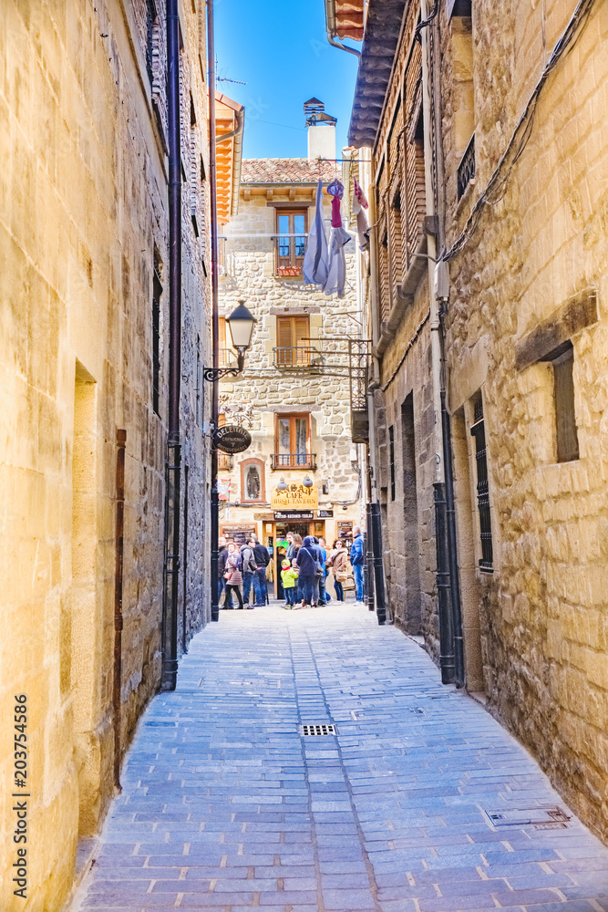 Laguardia, Alava, Spain. March 30, 2018: Narrow cobblestone street with facades of masonry stone houses and union with the main street of the town, full of people