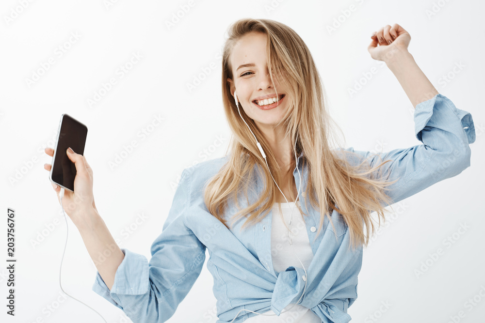 Come and dance with me. Portrait of joyful carefree european woman with fair hair, dancing while holding smartphone in raised hand and listening music in earphones, smiling broadly at camera Come and dance with me. Portrait of joyful carefree european woman with fair hair, dancing while holding smartphone in raised hand and listening music in earphones, smiling broadly at camera