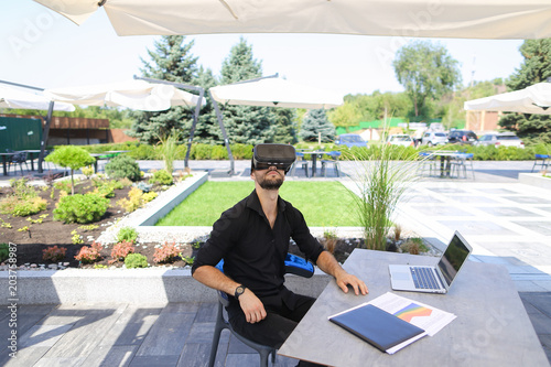 American businessman enjoying with virtual reality glasses, working with laptop and papers under beach umbrella near green plants. Concept of working on vacations and modern technology. photo