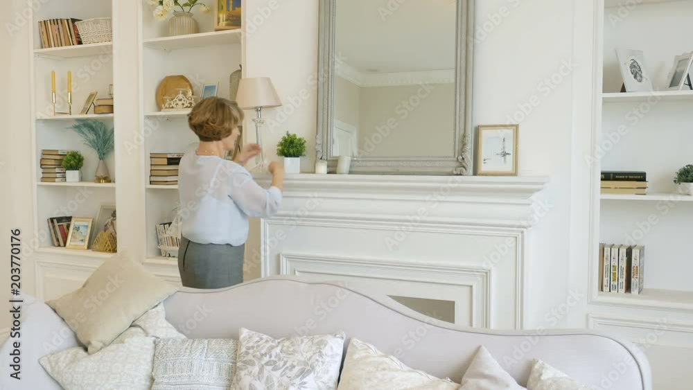 Senior woman in the living room wipes the dust, does the cleaning, looks in the mirror Senior woman in the living room wipes the dust, does the cleaning, looks in the mirror