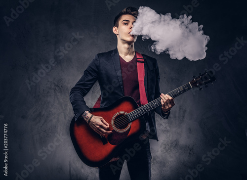 Handsome young musician with stylish hair in elegant clothes exhales smoke while playing acoustic guitar.