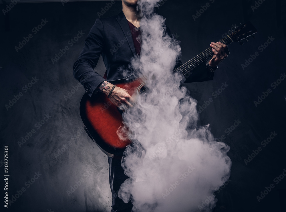 Stylish musician in elegant clothes exhales smoke while playing acoustic guitar.