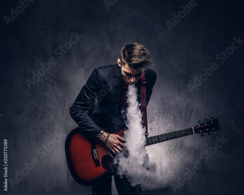 Handsome young musician with stylish hair in elegant clothes exhales smoke while playing acoustic guitar.
