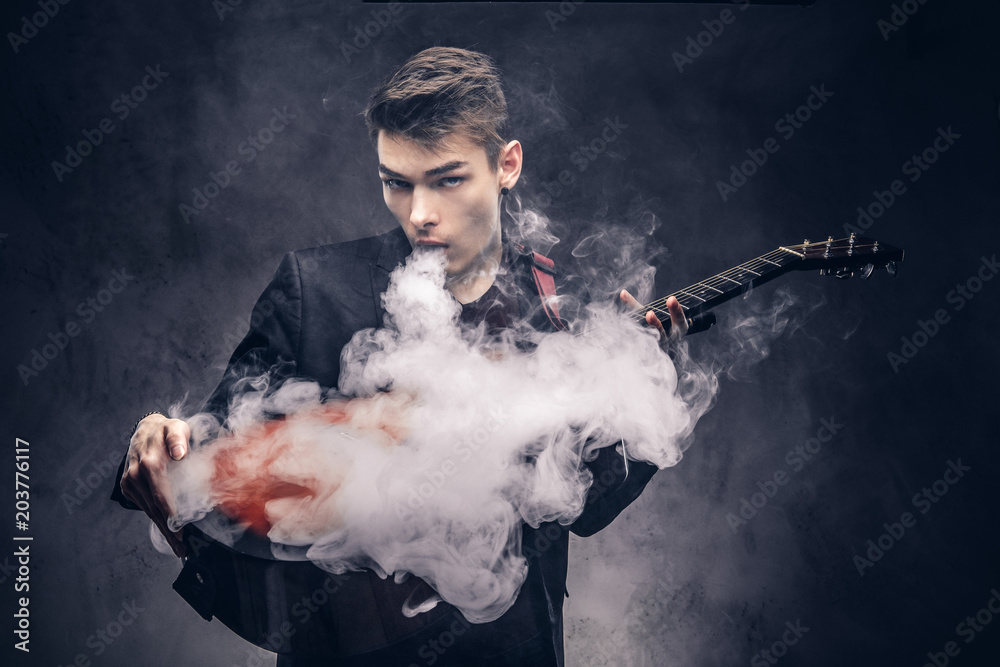 Handsome young musician with stylish hair in elegant clothes exhales smoke on his acoustic guitar.