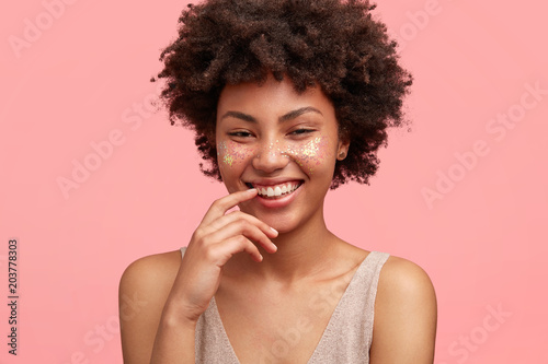 Close up shot of attractive young female wtith positive gentle smile, glad to have pleasant conversation with handsome guy, feels shy, isolated over pink background. Happy African American woman