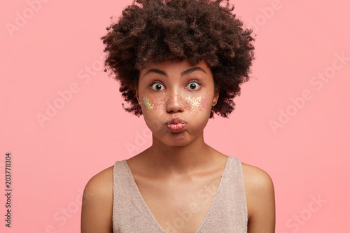 Funny amazed attractive African American female blows cheeks, has bated breath, cheeks decorated with spangles, stares at camera, prepared for party with friends, isolated over pink background