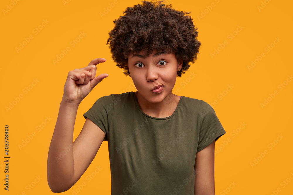 Studio shot of puzzled young cute African American female with curly crisp hair shows something tiny, demonstrates size of thing, wears casual t shirt, poses against bright yellow background. - obrazy, fototapety, plakaty 